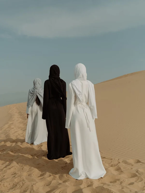 Illustrative: women in white and black abayas walking across sand dunes. Licensed stock photograph, not our own product.